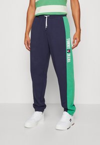 Marinblå och gröna joggers med dragsko i midjan och sidologga, med en mjuk bomullsblandning och avslutade med smalnande ärmar. Vita sneakers på fötterna.