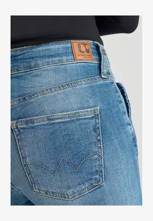 Blue denim jeans with a textured finish, featuring a leather brand patch and distinctive stitching on the back pockets.