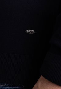 Granatowy dzianinowy sweter z prążkowymi mankietami i dołem oraz małą srebrną naszywką z logo. Gładka faktura i dopasowany fason.