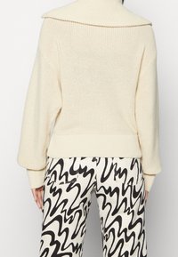 Maglione oversized di colore crema a maglia con un ampio colletto, polsini e orlo a coste; abbinato a pantaloni a righe black-and-white con linee ondulate.