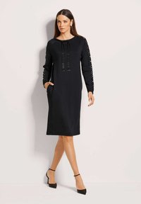 Frau in einem schwarzen knielangen Kleid mit paillettenbesetzten Ärmeln und Vorderdetails, schwarzen Knöchelriemenabsätzen, steht vor einem schlichten Hintergrund.