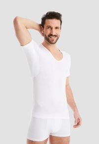 White short-sleeve shirt with a deep V-neck design and soft texture, paired with white briefs. Model is smiling, one hand above his head.