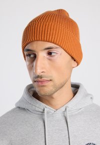 Bonnet côtelé en tricot orange, avec un sommet effilé et un ajustement serré. Porté avec un sweat à capuche gris, montrant une épaisseur et une texture moyennes.
