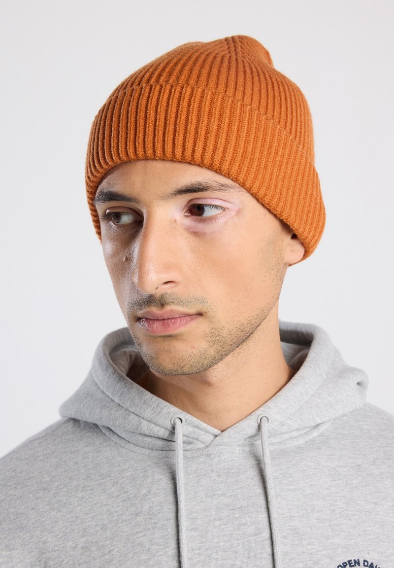 Bonnet côtelé en tricot orange, avec un sommet effilé et un ajustement serré. Porté avec un sweat à capuche gris, montrant une épaisseur et une texture moyennes.
