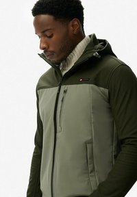 Donkergroene jas met grijze panelen, ritssluiting aan de voorkant en hoge kraag. Heeft een klein logo op de borst en een capuchon. Gladde, duurzame stof.