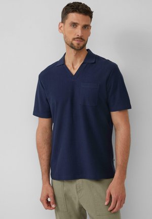 Marineblaues Kurzarmhemd mit V-Ausschnitt, einer Brusttasche, strukturiertem Stoff und einer lockeren Passform, getragen zu hellen Hosen.
