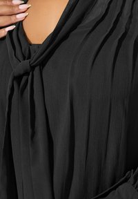 Camicia nera con una texture morbida e rigata, un dettaglio del collo annodato e un tessuto fluido, con maniche lunghe e leggero drappeggio.