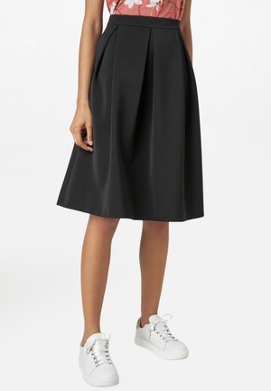 Femme portant une jupe plissée noire mi-longue, un haut à fleurs et des baskets blanches, debout devant un fond uni.