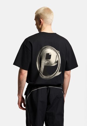 Blondyn z krótkimi włosami stoi tyłem, ma na sobie czarną koszulkę z dużym metalicznym literą P na plecach.