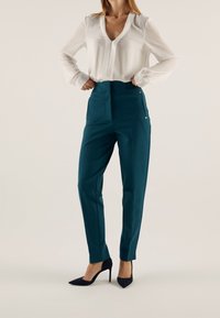 Pantalons taille haute en teal en tissu lisse, dotés de poches latérales avec des accents, associés à un blouse blanche et des escarpins noirs à bout pointu.