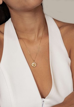 Woman wearing white halter top with gold necklace featuring a floral pendant resting above her chest against a neutral background.