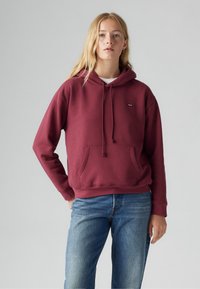 Burgundy hoodie made of soft fabric with a kangaroo pocket and drawstring hood. Paired with light blue denim jeans.