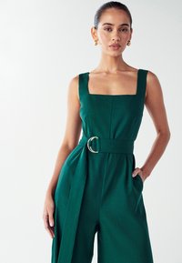 Mujer con el cabello pegado hacia atrás, vestida con un mono sin mangas de color verde con cinturón, escote cuadrado y pendientes de aro dorados, contra un fondo blanco.