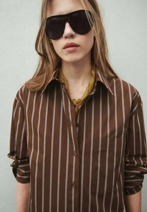 Mujer con cabello largo que lleva grandes gafas de sol negras, una camisa marrón a rayas con botones y un pañuelo amarillo en el cuello sobre un fondo liso.