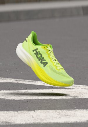 Scarpa da corsa Hoka gialla brillante e verde a mezz'aria sopra una strada con segnaletica bianca e piccoli detriti volanti sotto.
