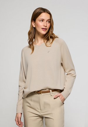 Beige oversized sweater with a V-neck and ribbed cuffs; features subtle logo embroidery and is paired with beige trousers and a brown belt.