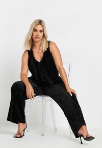 Svart sammetsjumpers med V-ringning, ärmlös design, med knappar fram och vida byxor. Modellen sitter på en genomskinlig plaststol.