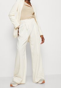 Costume à rayures blanches avec un pantalon large et un haut ajusté beige. Le tissu semble lisse, avec un subtil éclat et des détails de coupe décontractée.