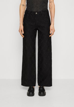 Persoon draagt zwarte wijd uitlopende gestructureerde broek, zwarte stevige schoenen en een mouwloos donkergrijs topje, staand tegen een effen achtergrond.