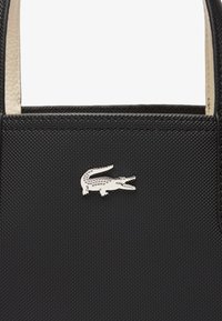 Sac à main noir texturé avec poignées crème, arborant un logo en crocodile argenté. Forme compacte avec un design structuré et sans quincaillerie visible.