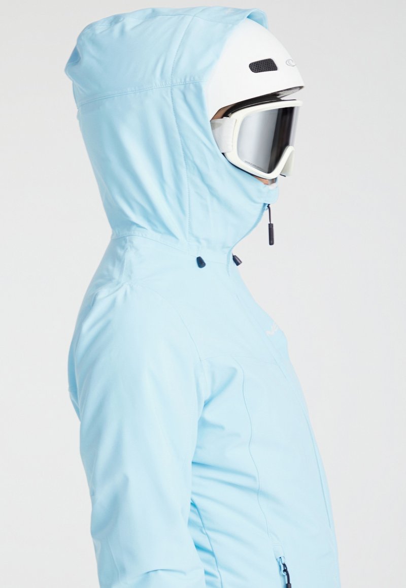 Giacca da sci blu chiaro con cappuccio, dotata di chiusura a zip e polsini regolabili, indossata con un casco da sci bianco e occhiali.