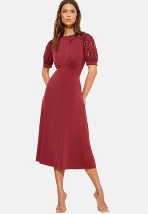 Femeie purtând o rochie burgundy până la genunchi cu mâneci bufante din dantelă, stând cu mâinile în buzunare și sandale bej cu toc înalt.