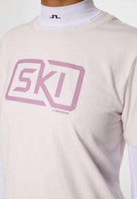 Jasnoróżowa koszulka wykonana z miękkiej bawełny, z dużą lawendową grafiką "SKI" oraz małym logo w dolnym rogu.