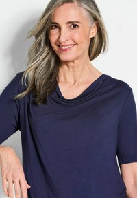 Marineblauw T-shirt met korte mouwen en een gedrapeerde col. Gemaakt van een gladde, lichte stof. Een vrouw met lang, steil grijs haar poseert met één hand op haar heup.