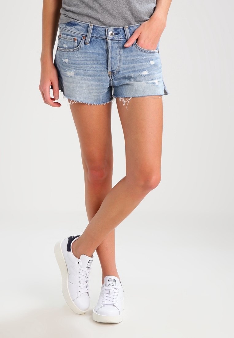 Persona che indossa pantaloncini di jeans azzurri con effetto usurato e sneaker bianche con dettaglio nero sul tallone, accavallando le gambe su uno sfondo semplice.