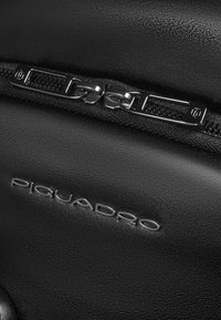 Zwarte leren tas met een gestructureerd oppervlak, voorzien van een metalen ritssluiting en hardware, en het ingeprinte logo "PIQUADRO" aan de zijkant.