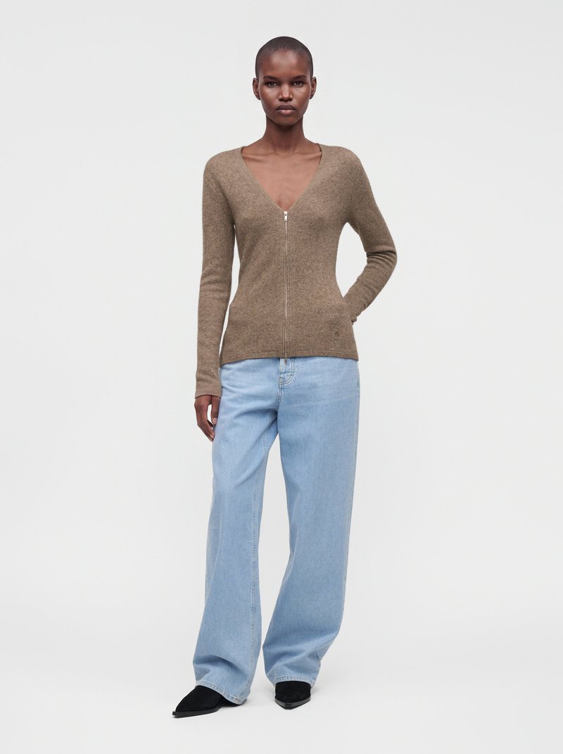 Personne mince portant un pull marron zippé, un jean bleu clair ample et des chaussures noires pointues, debout devant un fond blanc uni.