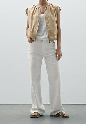 Personne portant un gilet beige sans manches zippé, un débardeur blanc, un pantalon blanc large et des sandales vert olive, debout devant un fond clair uni.