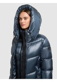 Marineblå pufferjakke med stor hætte, blank finish, quiltet design, lynlåslukning og justerbare snoretræk. Har mærkede detaljer.