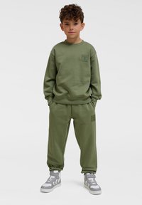 Felpa verde oliva e pantaloni abbinati realizzati in morbido tessuto, con polsini a costine e una piccola toppa con logo sul petto e sulla coscia.