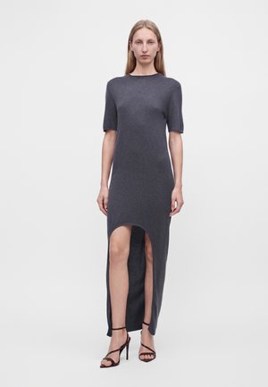 Femme portant une robe gris foncé à manches courtes, longueur décalée avec une fente sur le devant, et des sandales noires à talons hauts avec des brides, debout sur un fond uni.