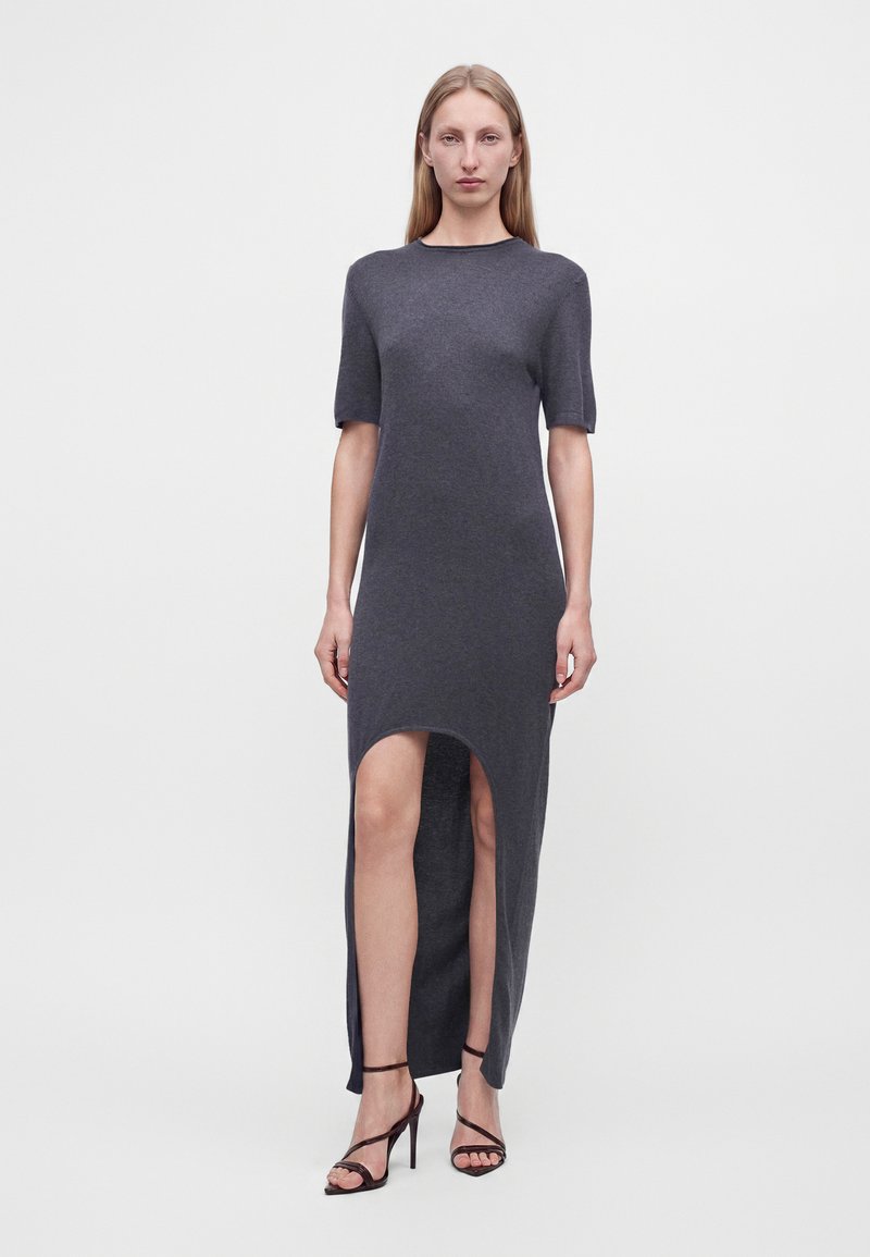 Femme portant une robe gris foncé à manches courtes, longueur décalée avec une fente sur le devant, et des sandales noires à talons hauts avec des brides, debout sur un fond uni.