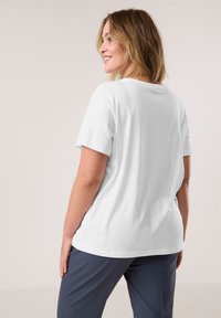 White cotton t-shirt with short sleeves, a round neckline, and a relaxed fit, paired with dark blue trousers, set against a neutral background.