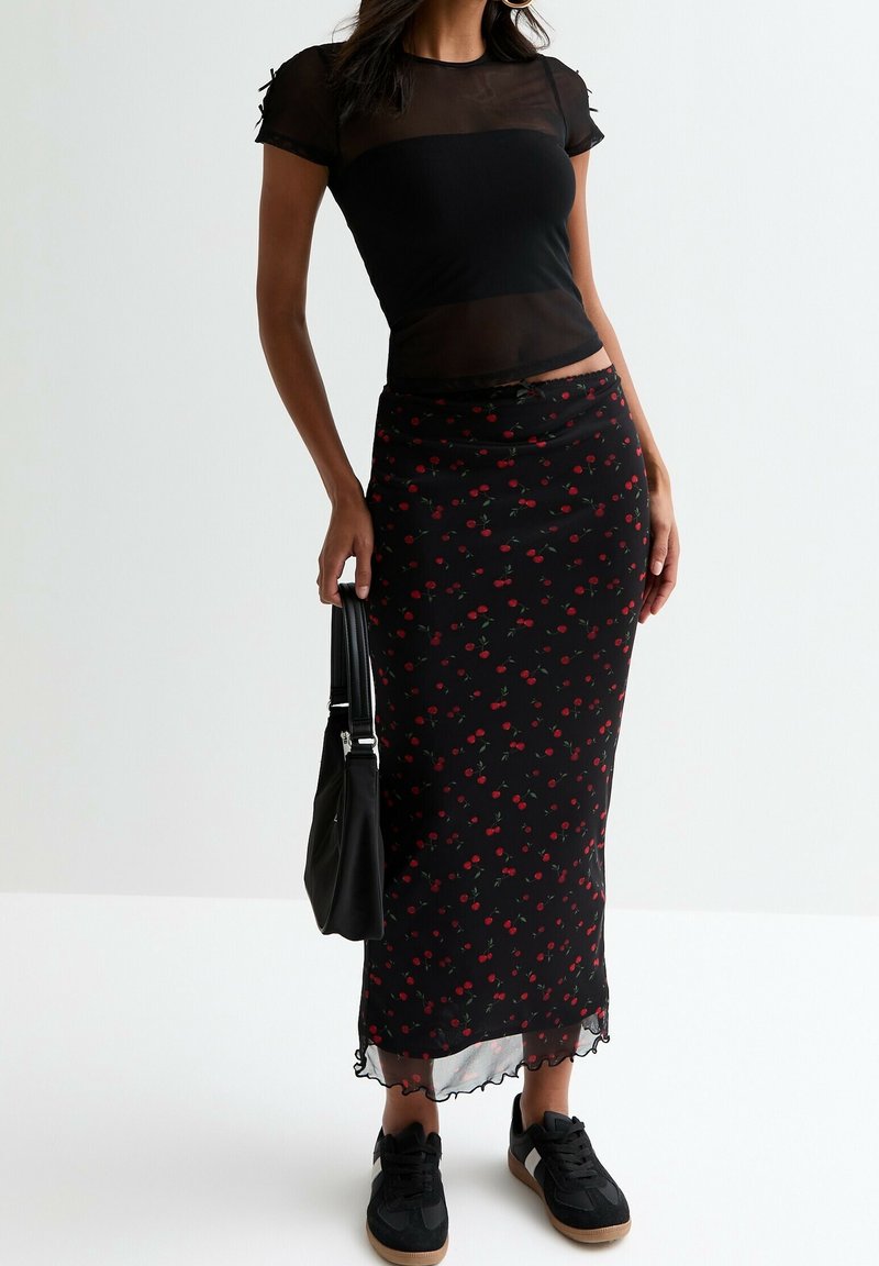 Femme portant un haut noir transparent à manches courtes, une jupe noire avec un petit imprimé cerise rouge, des baskets noires, et portant un sac à main noir.