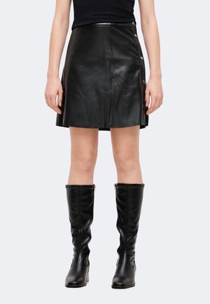 Mannequin portant une minijupe noire effet cuir avec des boutons sur le côté, un haut noir et des bottes noires jusqu’aux genoux.