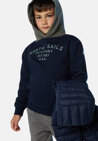 Sudadera con capucha azul marino con el texto "North Sails", bolsillo tipo canguro y puños acanalados, acompañada de una chaqueta acolchada azul marino doblada.