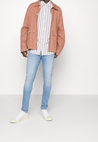 Chaqueta rosa claro con bolsillos delanteros sobre una camisa blanca y gris a rayas. Jeans skinny azul claro y zapatillas blancas completan el atuendo.