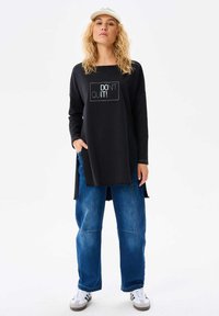 Czarna bluzka z długim rękawem z napisem "NIE PODDAWAJ SI!", luźny krój; zestawiona z luźnymi niebieskimi dżinsami i białymi trampkami, noszona z czapką.