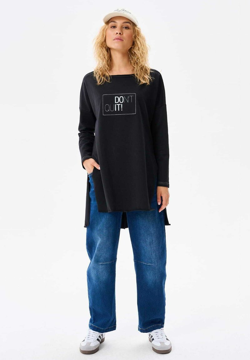 Czarna bluzka z długim rękawem z napisem "NIE PODDAWAJ SI!", luźny krój; zestawiona z luźnymi niebieskimi dżinsami i białymi trampkami, noszona z czapką.