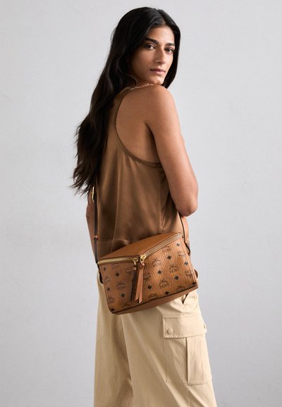 Tan leather crossbody bag with gold zipper and logo pattern, featuring a textured surface and a tassel accent.