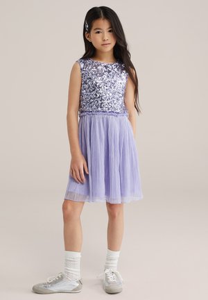 Levandų spalvos suknelė su blizgančiu liemenėliu ir klostuota sijonu, derinama su sidabriniais sportbačiais ir baltais tekstūruotais kojinėmis.