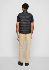 Czarna pikowana kamizelka z wysokim kołnierzem, z przeszyciami tworzącymi wzór. Nosi się ją na granatową koszulę, w połączeniu z beżowymi spodniami i czarnymi butami.