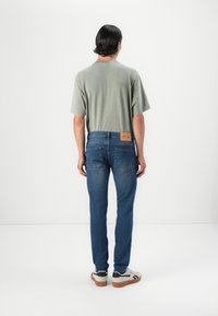 Camisa de manga corta gris claro, jeans de mezclilla azul y zapatillas blancas con detalles negros. La vista trasera muestra una etiqueta de cuero rectangular en los jeans.