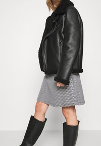 Mustat nahkatakki, jossa on karvakaulus, vetoketjukiinnitys ja sivutaskut. Yhdistetty kuvioituun hameeseen ja polven yli ulottuviin mustiin saapikkaisiin.