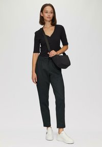 Schwarzes Kurzarmoberteil mit Knöpfen, kombiniert mit dunkelgrünen karierten Hosen und weißen Turnschuhen. Das Model trägt eine schwarze Umhängetasche.
