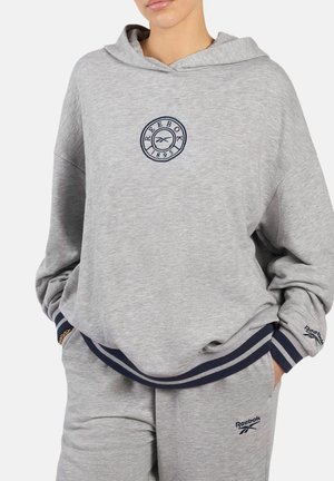 Personne portant un sweat à capuche Reebok gris clair et un pantalon de survêtement assorti avec un logo bleu marine et des bordures rayées, les mains dans les poches.